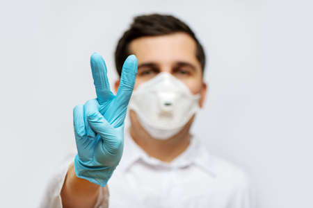 Portrait of doctor in medical uniform with a protective face mask and a gloved hand making a victory...