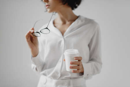 Cropped image of young woman holding eyeglasses and drinking coffee from paper cup over white background.の写真素材