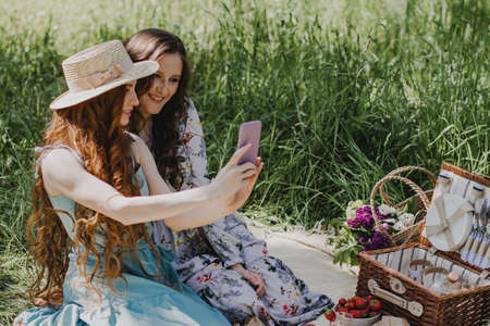 Two beautiful woman in summer dresses enjoying picnic outside and making selfie.の写真素材