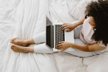 Young adorable African-American female sitting on the bed and typing on laptop.の写真素材