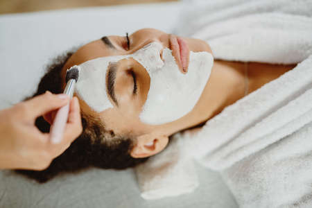 Beautician applies white clay face mask with a brush on a womans face.