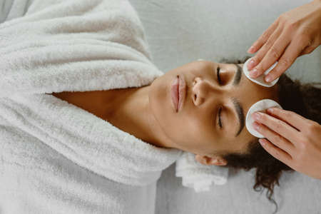 Professional beautician cleansing skin with cotton pads to dark-skinned young woman.