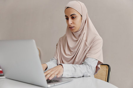 Young arabian woman in hijab sitting on her worklplace and working on laptop on light background.