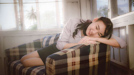 The Asian girl is smiling and siting in the couch, lying on sofa with light through the window at dawn vintage styleの写真素材