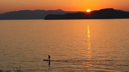A man is sailing on a board with an oar during a beautiful golden sunset over the water on Crete,...