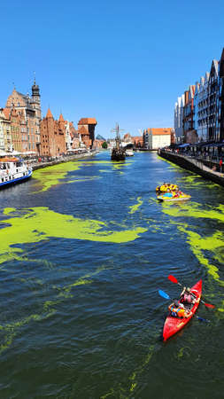 Gdansk, Poland - July 11, 2021: View of the old city of Gdansk on the Motlawa River. Tourists walk along the waterfront. On Motlawa green patches of duckweed. Middle of the river flows ship.のeditorial素材