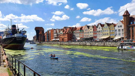 Gdansk, Poland - July 11, 2021: View of the old city of Gdansk on the Motlawa River. Tourists walk along the waterfront. On Motlawa green patches of duckweed. Middle of the river flows ship.のeditorial素材