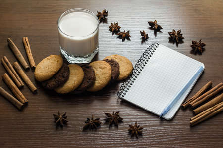 Flatley. A glass of milk, cookies, cinnamon and a notebook with a pen on a dark wooden table. White piece of paper with space for text. Diet and nutrition. Brown colorの写真素材