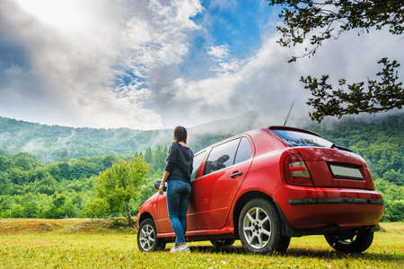 The girl stands near the red car looking at the mountains. Fog rises from the forest. The sky is...