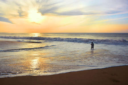 Young man walking on shore of the ocean with a camera in his hands against the rising sun. He nearly drowns in huge waves at high tide.の写真素材