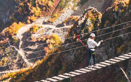 Unidentified girl in helmet walking along rope bridge hanging over abyss in mountains of Krasnaya...