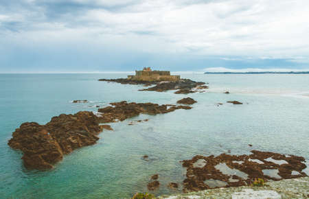Fort National, ancient fortress on tidal island Petit Be and rocks in turquoise water on cloudy day in Saint-Malo, Brittany, Franceの写真素材
