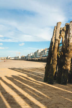 Wooden poles cast shadows on sandy beach and embankment with old and modern houses in background on sunny day in Saint-Malo, Brittany, Franceのeditorial素材