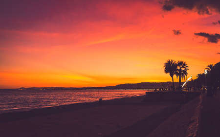 Pink, red and yellow colored sunset sky and silhouettes of people and palm trees at Promenade des Anglais in Nice, Cote dAzur, Franceの写真素材