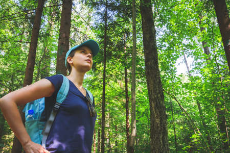 Young travelling girl with blue cap and backpack standing and looking up at trees in green forest on sunny summer dayの写真素材