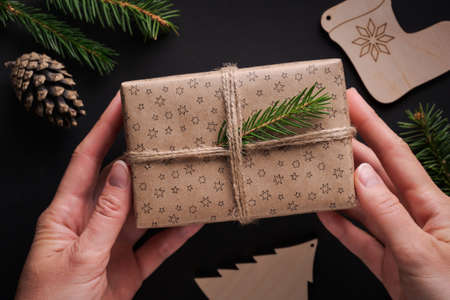 Female hands holding Christmas gift wrapped in craft paper and twine decorated with spruce twig...