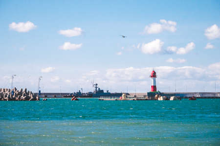 Seagull flying in blue sky above breakwater, moored ship, red and white lighthouse and turquoise sea