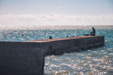 Silhouette of fisherman sitting alone with fishing rod on old pier and fishing at seaside on bright sunny dayの写真素材