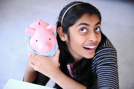 Cheerful, beautiful Indian girl hearing the sound of coins from Piggybankの写真素材