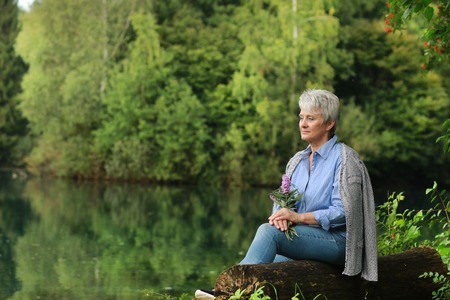 Serious senior woman sitting on a lake shore with flowers in her handsの写真素材