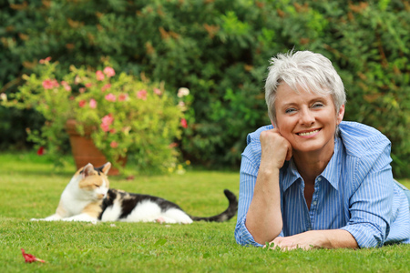 Happy senior woman is lying on the grass in the garden with a cat in backgroundの写真素材