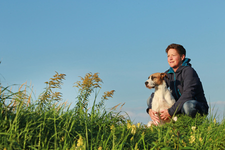 Happy woman with mixed breed dog sitting sideways in meadow against blue skyの写真素材
