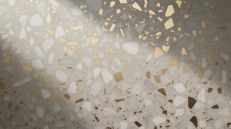 This image showcases a textured terrazzo surface with a mix of white marble, grey stone, and gold aggregate embedded in a light beige base. Sunlight casts dappled shadows across the surface, creating a dynamic interplay of light and shade. The pattern is irregular and organic, with varying sizes of chips.の素材