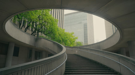 A concrete staircase curves upwards between textured walls, framed by a circular opening. Lush green trees contrast with distant glass skyscrapers under an overcast sky.の素材