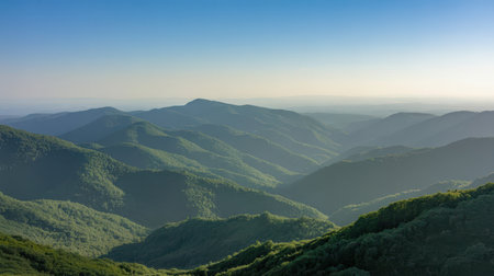 Layered green mountain ranges stretch to the horizon under a clear blue sky. Soft light of sunrise illuminates the rolling hills and valleys.の素材