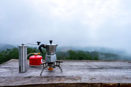 Portable coffee maker on fold-able gas stove with manual coffee grinder on wooden table in the middle of the forest with view of mountain, green trees and cloudy sky.の写真素材