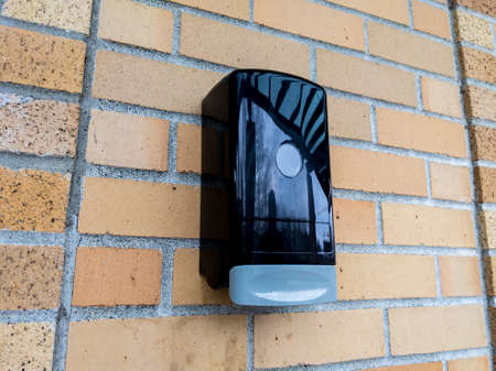 View of a black plastic hand sanitizer dispenser attached to a red brick wall on the outside of a buildingの写真素材