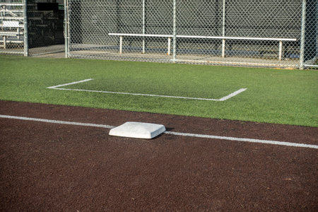 Angled view of a white base on a baseball field, with the view of a dugout in the background