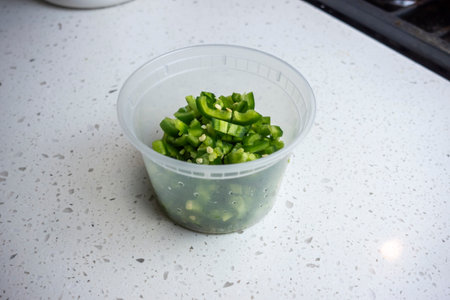 View of coloful diced jalapeno peppers in a plastic container on a kitchen counter, prepped for cookingの写真素材