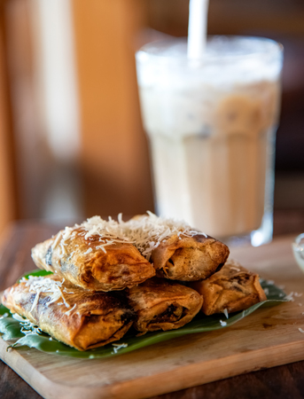 Fried banana  with cheese on the top of banana leaf and wooden cutting board. Selective focus with blur glass of coffee milk on background.の写真素材