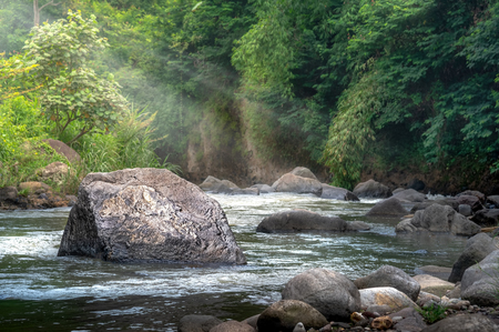 Deep Forest Stream Water Rushing by Big Rock in Middle River at Morning Sunshine and Warm Sunbeam or Sun Ray Through Tropical Forest Tree. Concept of Divine Harmony with Environmentの写真素材