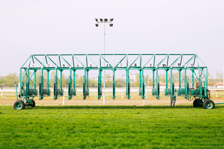 Empty horse racing starting stalls waiting for riders. Green colored starting gate on start by tractor machine at equestrian racehorse hippodrome. Outdoor sport racecourse competition equipmentの写真素材