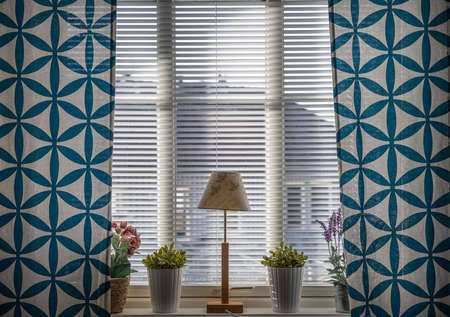A window with blue curtains, a lamp and flowers. Beautiful light comes in through the blinds