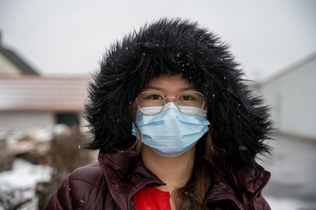 A teenage girl with in a winter jacket with a hood wearing a protective face mask. Picture in cold temperature and snowy conditionsの写真素材