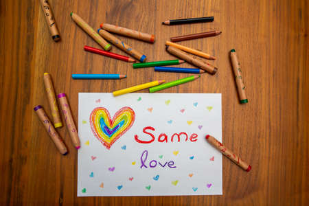 Child painting on a wooden table reading; "same love" with a rainbow heart, colorful crayons around itの写真素材