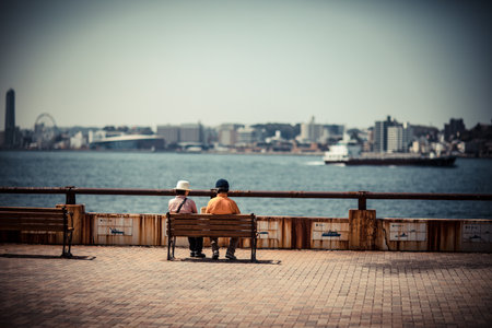 Two senior men sitting on a bench by the sea and looking at the cityの写真素材