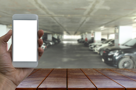 Empty wooden table space platform and Hand holding smartphone with white blank screen over blurred  Parking lot in the mall background for product display montage.の写真素材