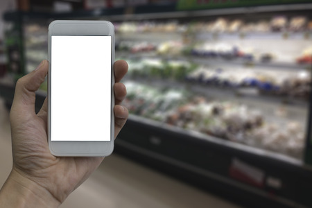 Hand holding smartphone with white blank screen over blurred supermarket and retail store in...