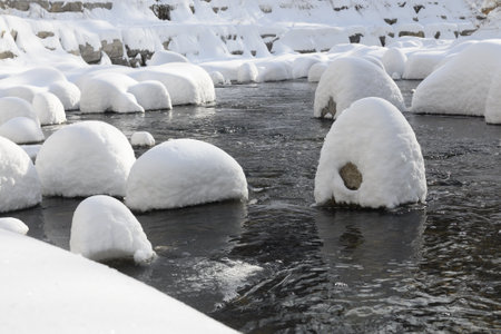 Large rocks covered in thick snow sit in a flowing creek, creating a unique winter landscape with soft, rounded shapes.の写真素材