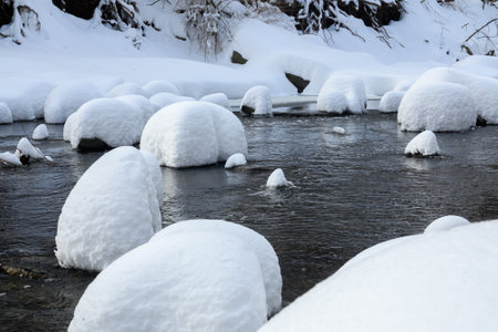 Large rocks covered in thick snow sit in a flowing creek, creating a unique winter landscape with soft, rounded shapes.の写真素材