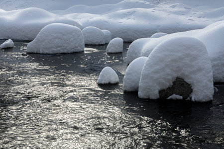 Large rocks covered in thick snow sit in a flowing creek, creating a unique winter landscape with soft, rounded shapes.の写真素材
