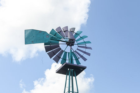Vintage metal windmill stands tall against a blue sky with white clouds and trees in the background.の写真素材