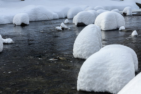 Large rocks covered in thick snow sit in a flowing creek, creating a unique winter landscape with soft, rounded shapes.の写真素材