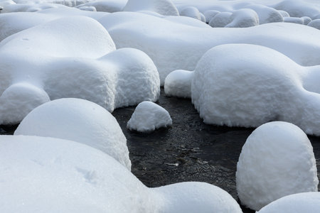 Large rocks covered in thick snow sit in a flowing creek, creating a unique winter landscape with soft, rounded shapes.の写真素材