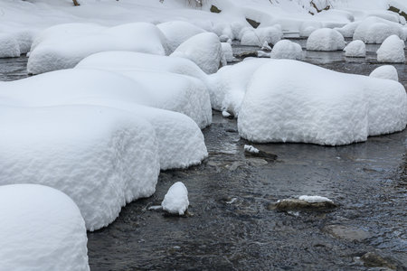 Large rocks covered in thick snow sit in a flowing creek, creating a unique winter landscape with soft, rounded shapes.の写真素材