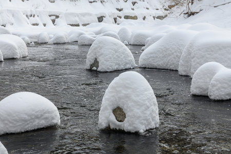 Large rocks covered in thick snow sit in a flowing creek, creating a unique winter landscape with soft, rounded shapes.の写真素材